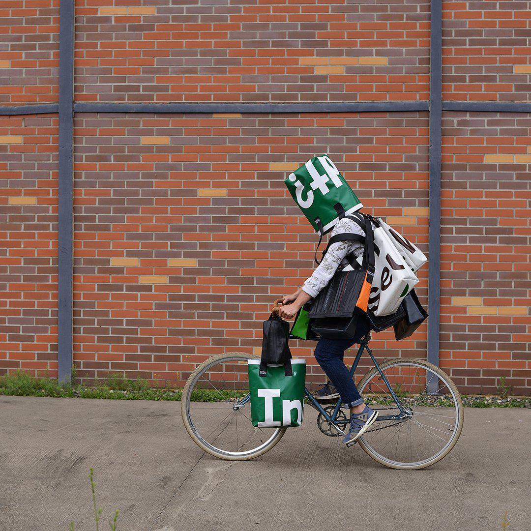 Person auf dem Fahrrad vor einer Backsteinwand, bepackt mit grünen Taschen aus bedruckter LKW-Plane.