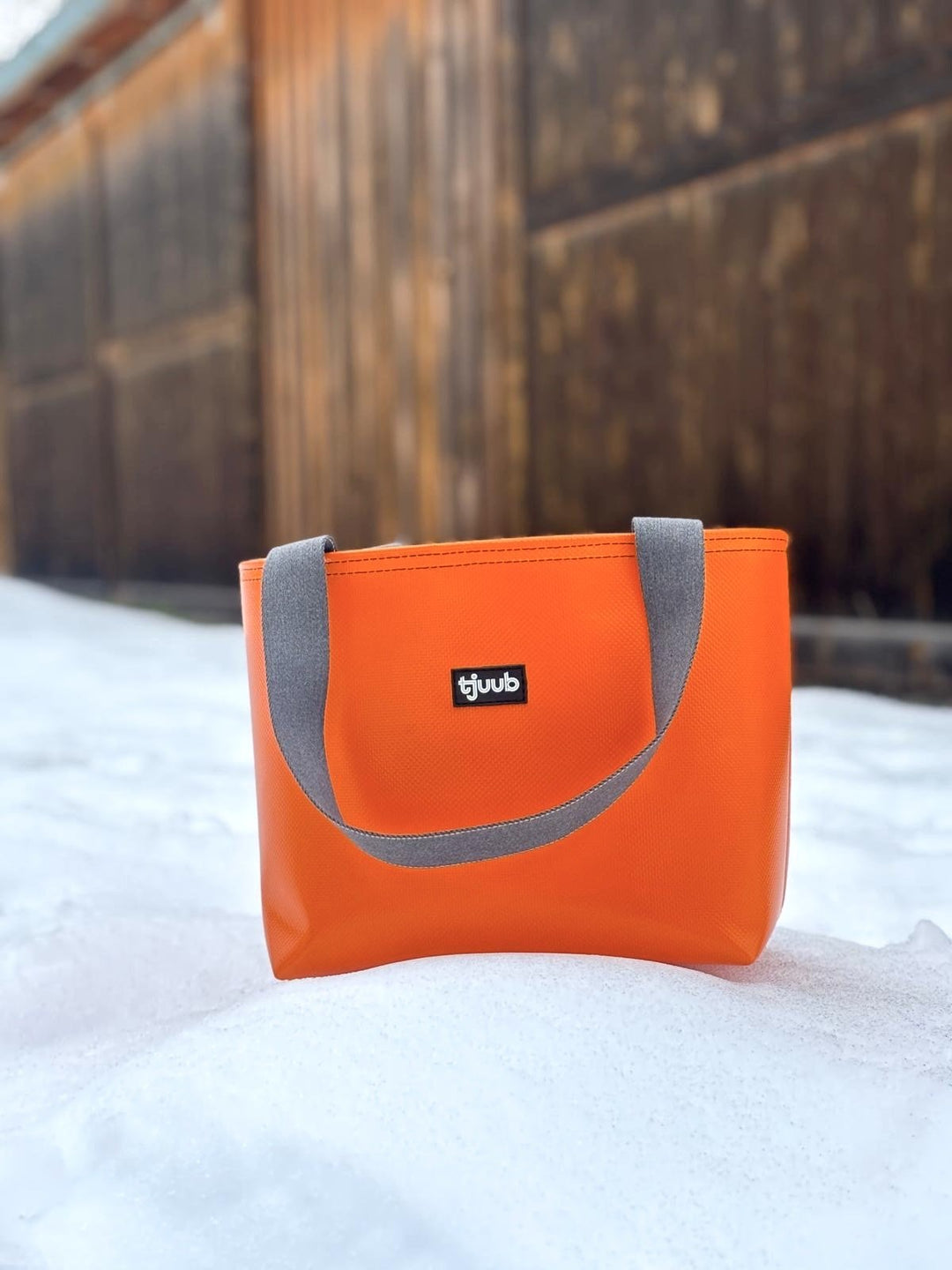 Orange Tasche Sila ohne Reißverschluss aus LKW-Plane steht im Schnee vor einer Holzwand.