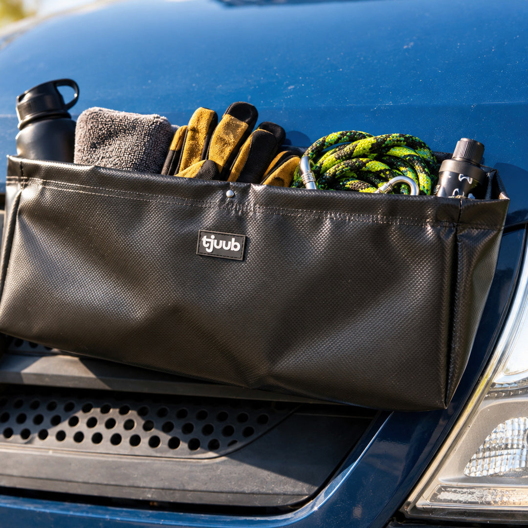 Muddus Organizer haftet magnetisch außen an der Motorhaube eines blauen Vans, darin Trinkflasche, Tuch, Handschuhe und Hundeleine.