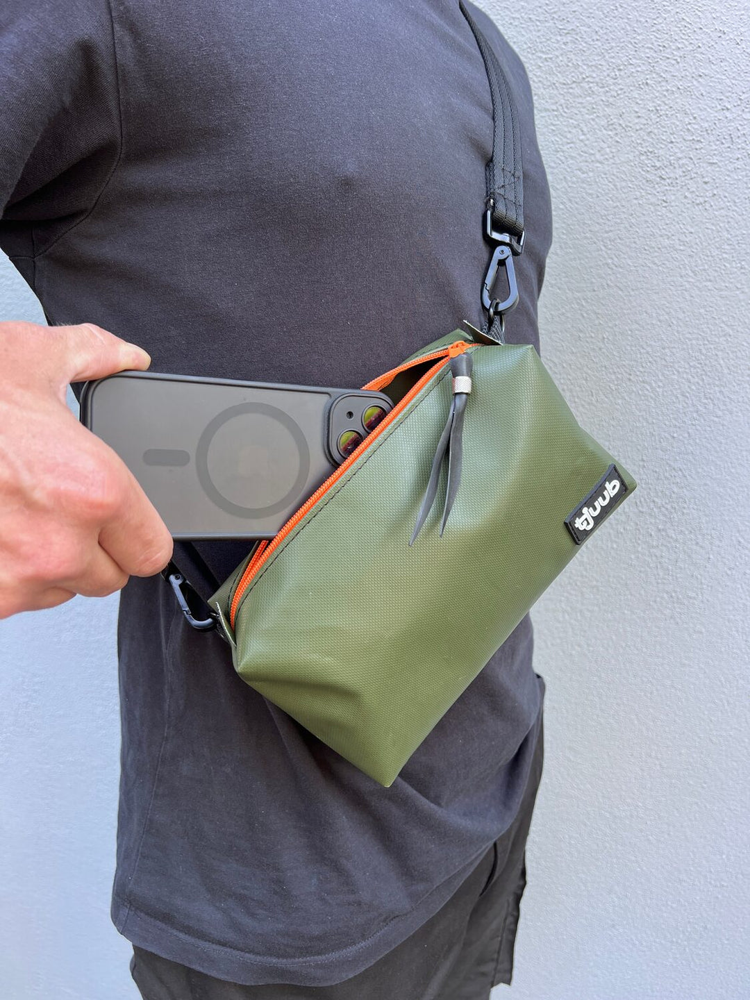 Olivgrüne Umhängetasche Una aus LKW-Plane an der Hüfte getragen, daneben ein Smartphone – passt als Handytasche.